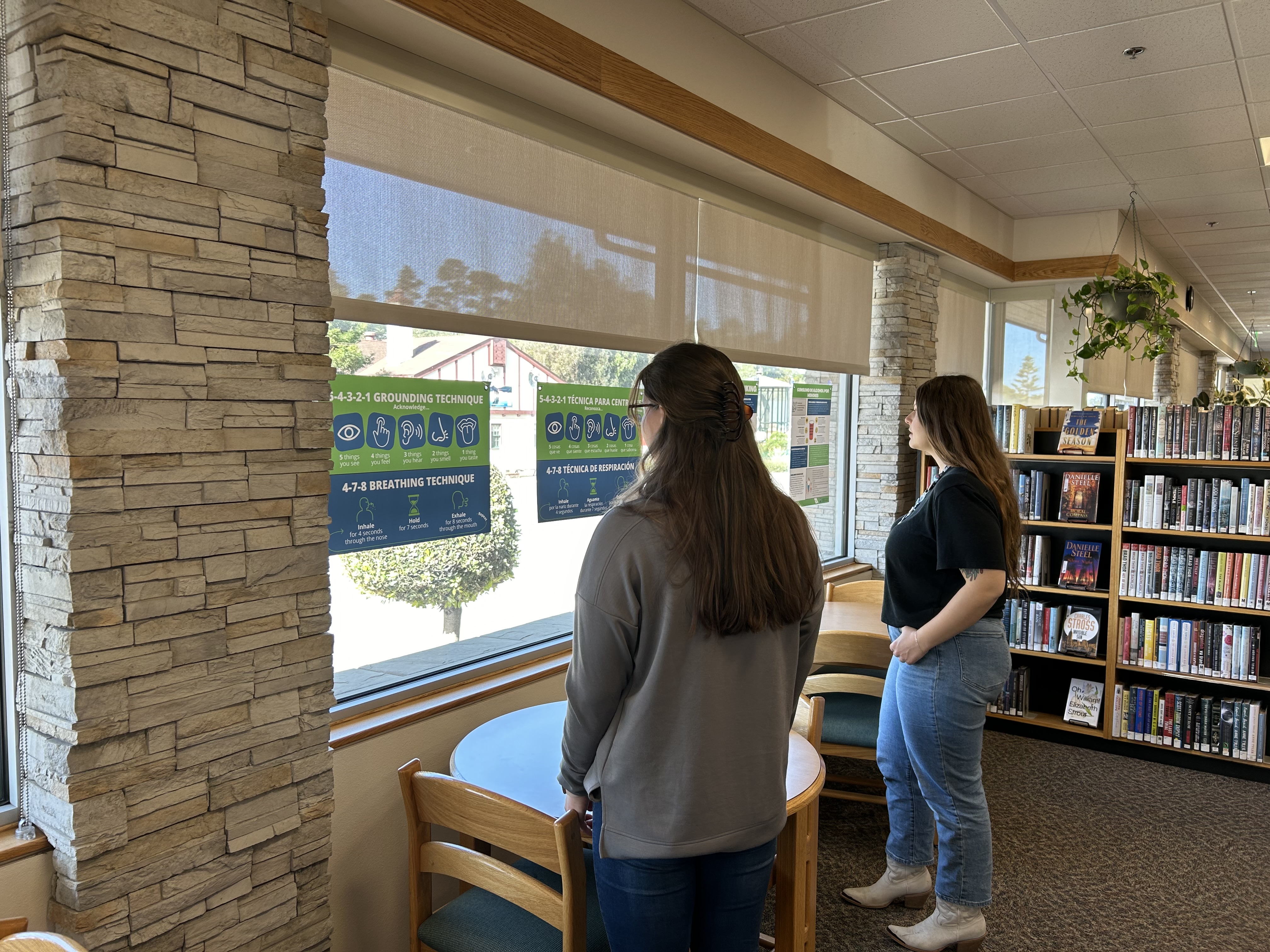 Two women are standing by a window, reading the posters. Click to view article, Community Libraries Host Gallery to Spotlight Mental Wellness and Substance Use Awareness