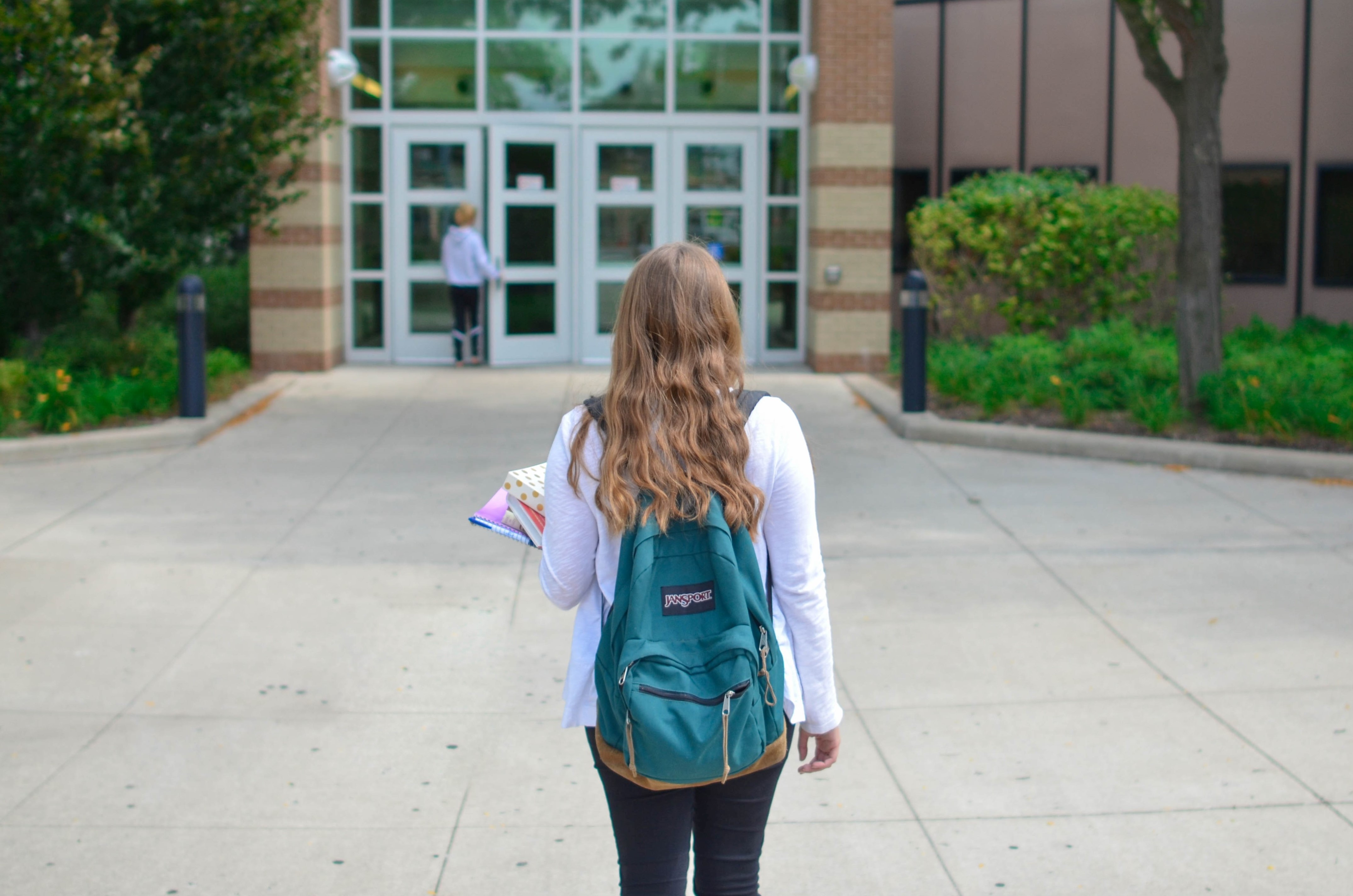 Student wearing teal backpack and holding books stands in front of school entrance