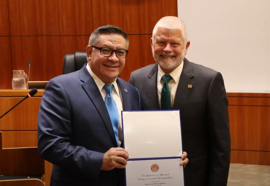 Image of Congressman Salud Carbajal and Supervisor Bruce Gibson in the Board of Supervisor chambers. Congressman Salud Carbajal holds a certificate of white paper which has a recognition. 