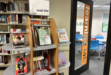 Image of books on display on shelves in the library. The shelf has a sign that reads "Read on!" 