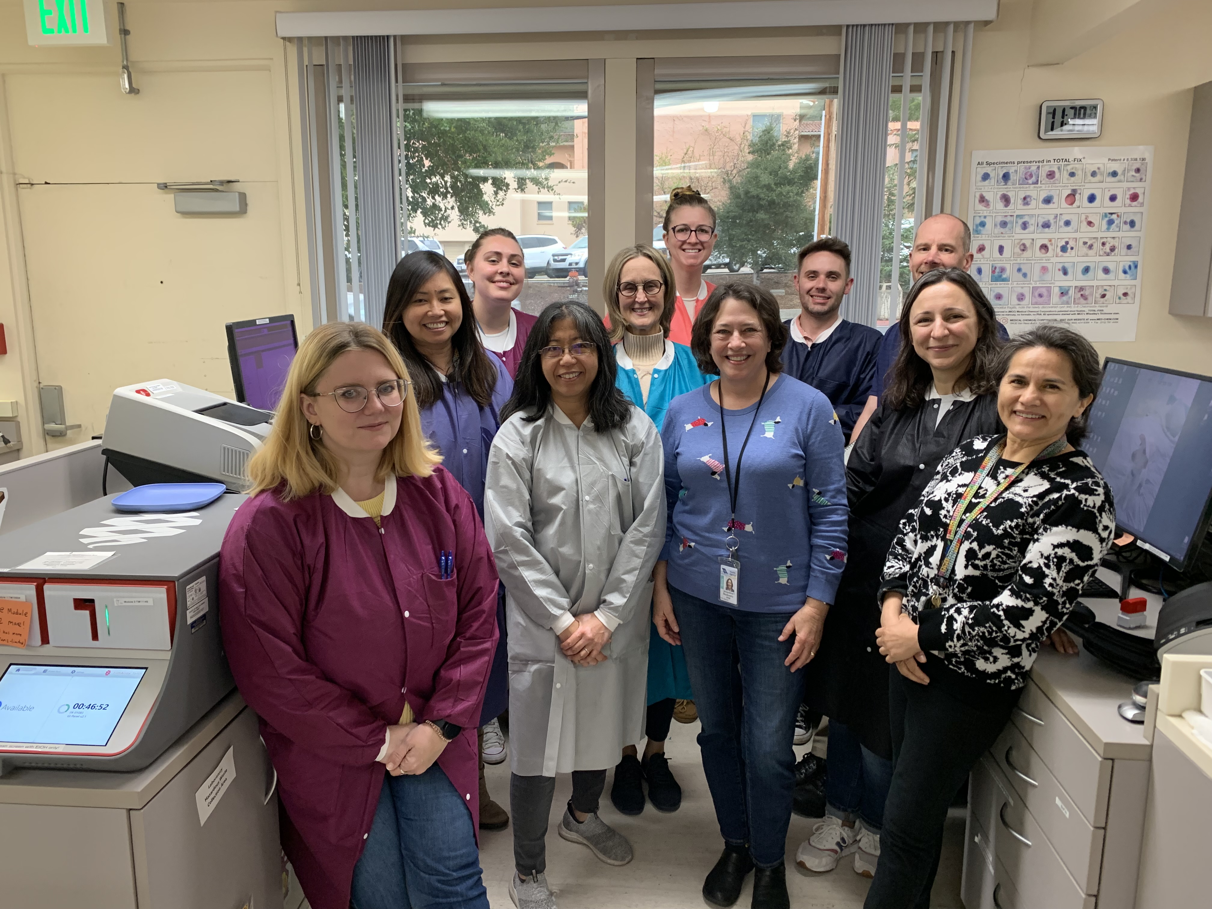 Team of laboratory scientists wearing gowns smiling in a group amid machinery.
