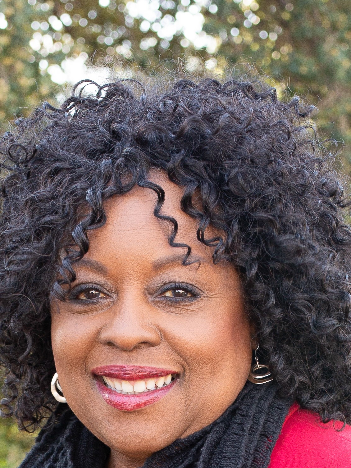 Image of Cheryl Vines with a red top and a black scarf around her neck. In the background there is a tree with green leaves