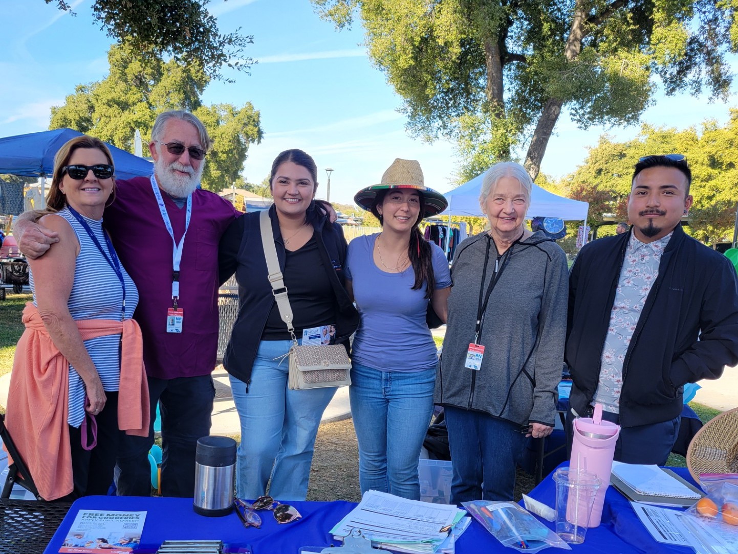 Six people smiling, standing together out under oak trees behind a table with a blue table cloth.