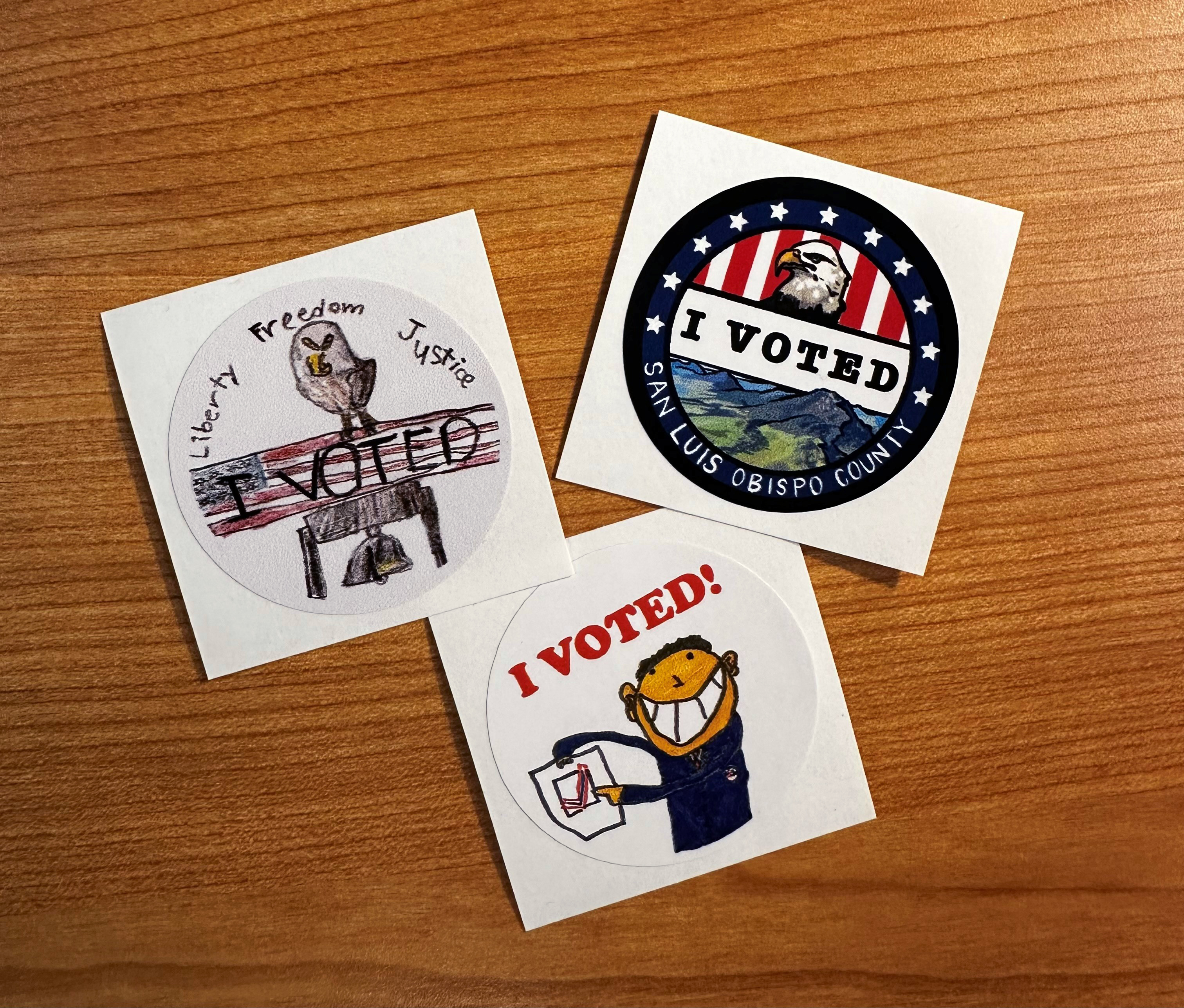 Photo of three hand-drawn stickers that each say I Voted. The feature bald eagles, rolling hills, and a smiling person pointing to his ballot.