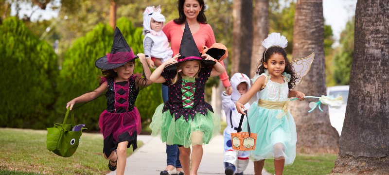 Children in costumes run on a sidewalk in front of an adult caregiver