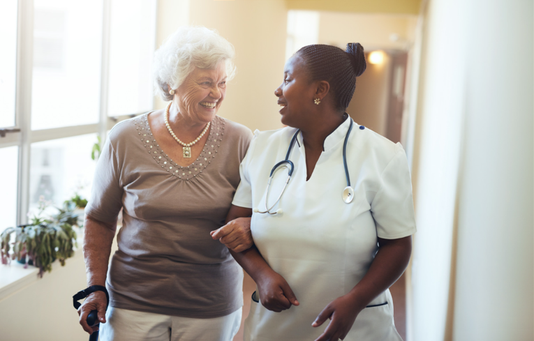 Older woman with short while walking down a sunlit hallway arm in arm with a caregiver chatting with her.